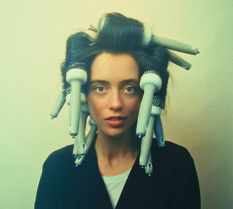 Portrait of a person in heated hair rollers against a pale wall, wearing a dark cardigan.