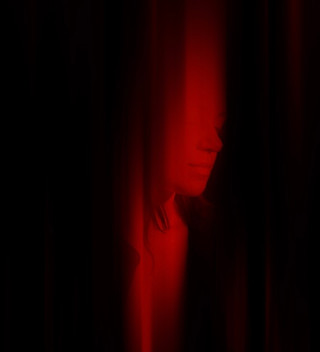A face half hidden behind a dark curtain, lit by a narrow slit of red light.