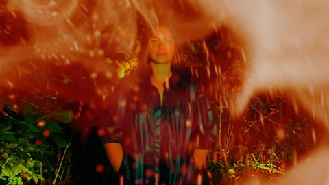 Portrait shot through a blur of orange firelight and leaves, the subject centred in a garden at dusk.