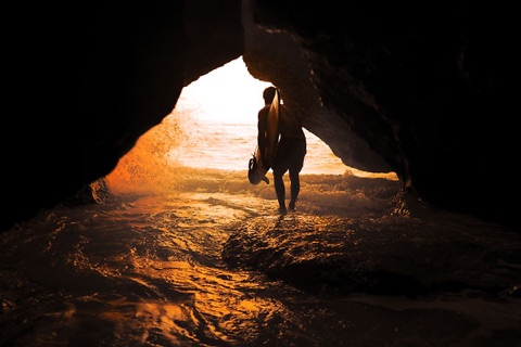 Silhouette of a person carrying their shoes, walking out of a sea cave into the surf at sunset.