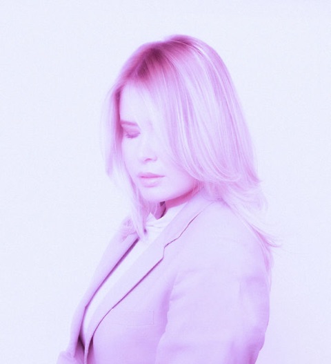 Profile portrait of a person with long blonde hair in a pale suit, washed in monochrome pink.